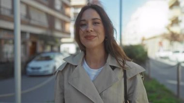 Güneşli şehir caddesinde hava öpücüğü veren bir kadın bej renkli trençkotlu, neşeli bir ifade ve şehir arka planına sahip, açık hava sıcaklığını kucaklayan ve canlı bir gülümseme takınıyor..