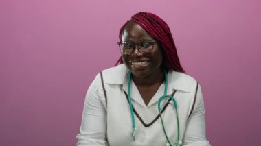 Gözlüklü ve steteskoplu Afro-Amerikalı kadın izole edilmiş pembe arka plan duvarına güvenle gülümserken....