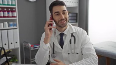 Steteskoplu beyaz önlüklü genç bir doktor hastane ofisinde akıllı bir telefonla konuşurken gülümsüyor..
