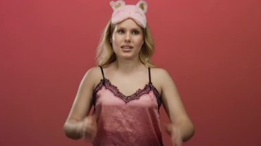 Uyku maskeli pembe gecelikli bir kadın, elleri havada ve saçlarını taramış bir rahatlama ve neşe karışımı sergiliyor, canlı kırmızı bir zemin üzerinde şakacı bir şekilde poz veriyor..