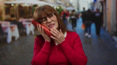 Dişi ağrıyan İspanyol son sınıf öğrencisi kadın, şehir restoranının terasında gözlük ve kırmızı süveter giyerek otururken yanağında buz torbası tutuyor..