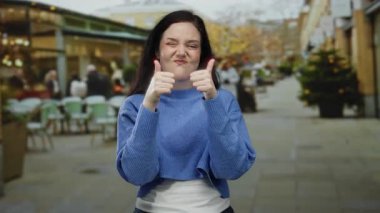 Bir restoranın dışındaki canlı bir teras caddesinde jest yapan, mavi kazak giyen, insanlarla ve bitkilerle çevrili, canlı ve çekici bir şehir manzarası yaratan bir kadın..