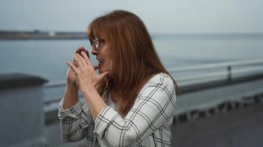 Gözlüklü ve kareli tişörtlü yaşlı İspanyol kadın deniz kenarında bir gezinti alanında bağırarak arka planda okyanusla birlikte açık hava heyecanını yakalıyor..