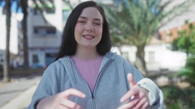 Güneşli bir sokakta palmiye ağaçlarıyla gülümseyen bir kadın, açık havada ellerle kalp oluşturan, sıradan bir kapüşonlu giyen bir kadın..