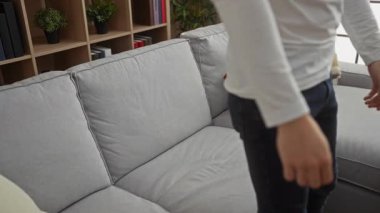 Genç bir adam şık bir oturma odasındaki koltukta rahat bir şekilde dinleniyor. Arka planda renkli yastıklar ve raflar var. Kapalı alanda rahat bir atmosfer sergiliyor..