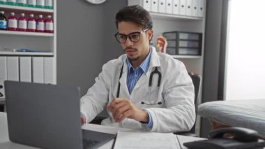 Klinikteki genç erkek doktor dizüstü bilgisayarla çalışıyor ve telefona odaklanmış bir ifadeyle cevap veriyor..