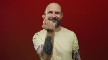 Kel, sakallı, parlak kırmızı bir duvara karşı kalp hareketi yapan, sıradan, dostane bir ifade sergileyen, kel ve dövmeli bir adam..