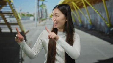 Beyaz elbiseli, uzun saçlı genç bir kadın güneşli bir şehir sokağında neşeli bir şekilde dışarıyı işaret ederek şehir mimarisini ve canlı bir şehir atmosferini ön plana çıkarıyor..