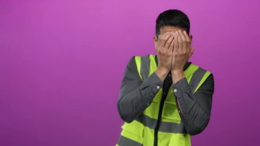 Güvenli yelek giyen genç adam, elleri parlak pembe bir arkaplanda, mizah ve şaşkınlık ifadesi sergiliyor..