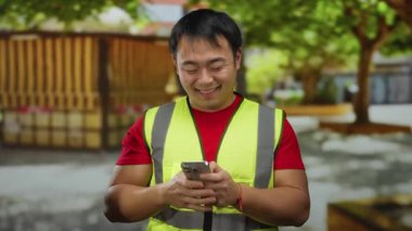 Parkta gülümseyen bir adam açık havada sarı yelek giyip akıllı telefondan mesajlaşırken iletişim ve aktiviteyi temsil ediyor..