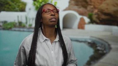 Gözlüklü bir kadın havuz başında güneşli bir günün tadını çıkarıyor, rahatlama ve rahatlama çağrıştırıyor, yemyeşil ve şık mimariyle çevrili, mükemmel bir kaçış için..