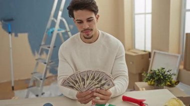 Yeni boyanmış merdiven ve kutularla dolu bir odada bize dolar tutan genç adam, mutlu bir ifade ve günlük giysiler gösteriyor, ev yenileme sırasında mali başarı öneriyor..