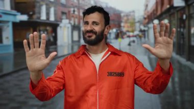 Kırmızı hapishane tulumlu, sakallı İspanyol adam yağmurlu bir sokakta elleri havada duruyor..