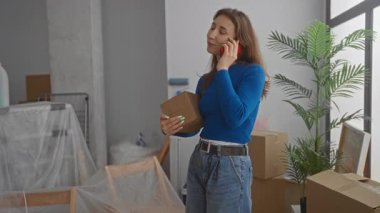 Yeni dairede kutular ve saksılarla çevrili bir kadın telefonla konuşuyor. Evin içini gösteriyor. Oturma odasında taşınıyor..