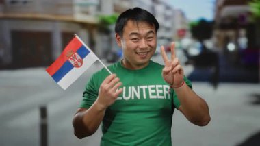 Kentsel caddede gönüllü tişört giyen genç adam barış işareti gösteriyor ve gülümsüyor, kentteki kültürel çeşitliliği simgeliyor..