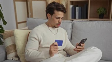 Oturma odasındaki kanepede oturan genç adam elinde kredi kartı ve akıllı bir telefonla online alışverişe odaklanmış sıradan bir kazak giyiyor..