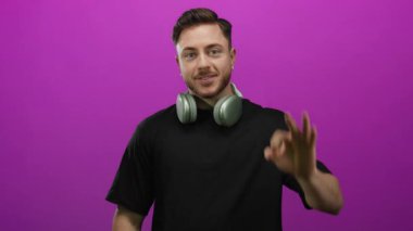 Kulaklıklı genç adam gülümsüyor ve parlak pembe bir arka plana el hareketi yapıyor. Kendine güveni ve pozitifliği var..