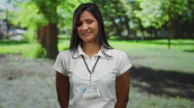 Beyaz gömlekli, isim rozetli, yeşil ağaçlar ve çimlerle çevrili genç Latin bir kadın..