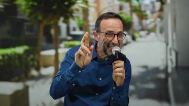 Şehir parkında mikrofona konuşan bir adam, arka planda ağaçlar ve binalar, açık havada gözlüklü ve mavi gömlekli, orta yaşlı, anlamlı el hareketleriyle.