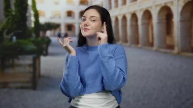 Mavi kazaklı kadın üniversite kampüsünde kendinden emin bir şekilde duruyor ve tarihi yapı geçmişi olan bir fikir jesti yapıyor..