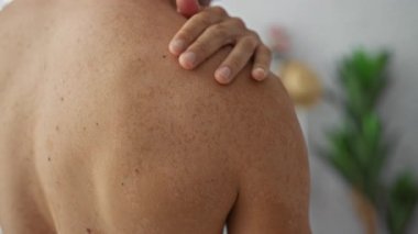 İçerideki genç adam, rahat bir ev ortamında çıplak sırt ve elle omzuna dokunuyor. Etrafı hafif ışıklandırma ve sıcak bir iç atmosferle çevrili..