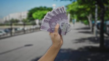 Endonezya Rupiah 'ı elinde tutan adam güneşli bir şehir caddesinde, canlı doğal geçmişi, kentsel çevresi ve para birimi detaylarıyla finansal konsepti vurguluyor..