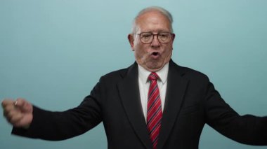 Takım elbiseli kıdemli işadamı mavi arka plana karşı güçlü bir jest yapıyor. Başarıyı ve güveni simgeliyor..