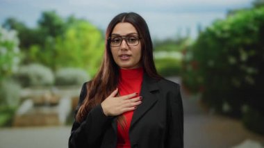 Kırmızı balıkçı yaka kazaklı ve gözlüklü siyah ceketli kadın yemyeşil parkta mahkumiyetle konuşuyor..