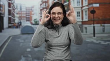 Esmer saçlı, gülümseyen ve el kol hareketi yapan bir kadın. Arka planda kırmızı tuğlalı binalar olan bir şehir caddesinde..