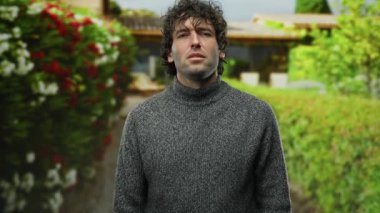 Hispanik bir adam parkta canlı çiçeklerin ve yeşilliğin bulanık arka planına karşı hayal kırıklığını dile getiriyor, doğal çevrede duygusal olarak samimi bir portre çekiyor..