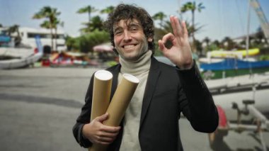 Elinde evrak tüpleriyle gülümseyen adam deniz kıyısında arka planda tekneler ve güneşli gökyüzünün altında palmiye ağaçları olduğunu gösteriyor.