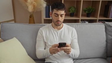 Oturma odasında bir koltukta oturan genç adam düşünceli bir ifadeyle telefona bakıyor, günlük beyaz bir gömlek giyiyor ve modern bir ev dekorasyonu sergiliyor..