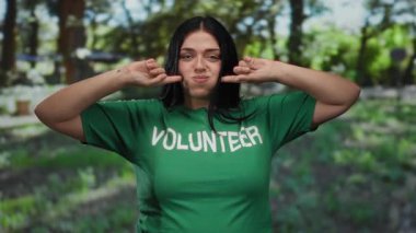 Artı, yeşil tişörtlü, parkta gönüllü bir kadın, açık hava aktivitelerine neşe içinde katılıyor, ağaçların ve çimenlerin arka planında güneş ışığının altında..