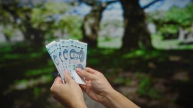 Arka planda ağaçların bulanıklaştığı bir şehir parkında Çin Yuan banknotlarıyla el ele tutuşmak mali ya da ticari bir alışveriş sahnesi izlenimi uyandırıyor..
