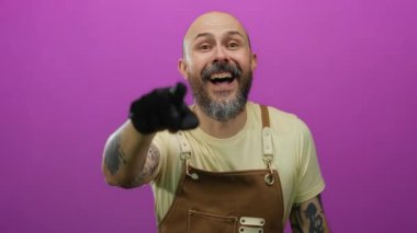 Sakallı kel adam gülüyor ve işaret ediyor. Önlük ve eldiven takıyor. Pembe arka planda..