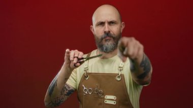 Elinde makas tutan sakallı kel bir adam canlı kırmızı arka plan dövmelerine ve odaklanmış ifadeye karşı önlükle güvenle poz veriyor..