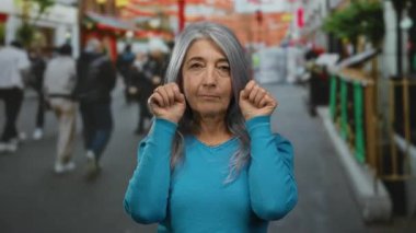 Kıdemli, gri saçlı, mavi kazaklı bir kadın arka planda bulanık yayalarla kalabalık bir sokakta şakacı hareketlerle hayat dolu bir şehir manzarasını yakalıyor..