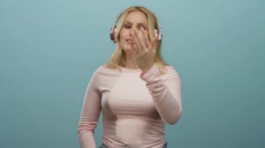 Sarı saçlı ve kulaklıklı bir kadın, mavi arka planda jestler yapıyor, hoş bir ifadeyle pembe bir bluz giymiş, hoş bir karşılama havası veriyor..