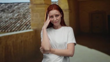 Açık üniversite avlusunda duran kadın, el ele tutuşmuş, düşünceli ve düşünceli görünüyor, sakin ve resimli bir akademik ortam sergiliyor..