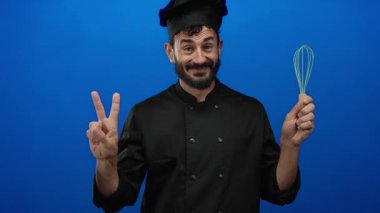 İspanyol aşçı şefin elinde çırpı jestleri var. Mutfak stüdyosunda mavi arka planda barış işareti var..