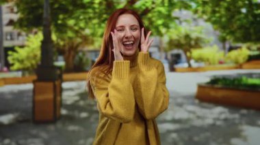 Sıcak bir kazakla gülümseyen bir kadın, canlı bir parkta güneşli bir günün tadını çıkarıyor, yemyeşil ağaçlar ve yeşilliklerle çevrili, bir anlık neşe ve doğal güzelliği yakalıyor..