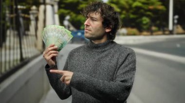 Şehir ortamında elinde Rus rublesi tutan bir adam yan tarafı gösteriyor, süveter giyiyor ve merakını ve dikkatini gösteriyor..