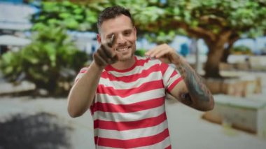 Genç adam gülümsüyor ve güneşli bir günde çizgili bir gömlek giyerken şehir ortamında başparmağıyla işaret ediyor.