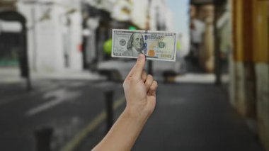 Adam bizi bir şehir caddesinde tutuyor, açık bir şehir ortamında yüz dolarlık banknotlar sergiliyor, finans ve ekonomik değişimin özünü yakalıyor..