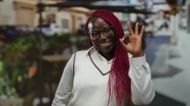 Kırmızı örgülü, gülümseyen ve el kol hareketi yapan bir kadın. Canlı bir şehir caddesinde, restoranın terasında, gün içinde..