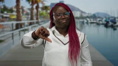 Örgü örgülü bir kadın açık havada açık bir gökyüzünün altında botları ve palmiyeleri olan bir limanda baş parmağını sallıyor..