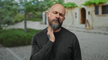 Sakallı kel bir adam şehir ortamında boğaz ağrısı çekiyor. Gün ışığında ağaçlar ve taş binalarla dolu bir şehir zemininde rahatsızlık gösteriyor..