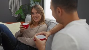 Çift, sıcak bir oturma odasında, kırmızı kupalarla kahvenin keyfini çıkarıyor, sıcak bir gülümseme ve rahat ve samimi bir atmosfer yaratıyor..