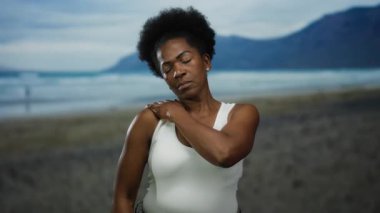 Sahilde omzunu acıyla tutan bir kadın dingin bir okyanus zeminine yaslanmış, plajın keyfini çıkarırken kolsuz bir bluzla eğleniyor..