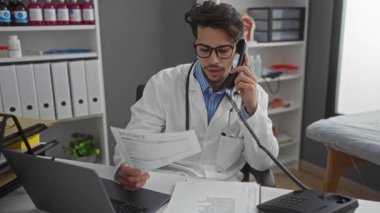 Klinikteki genç erkek doktor, onu çevreleyen belgeler, bilgisayarlar ve tıbbi dosyalarla telefonda konuşurken belge okuyor..
